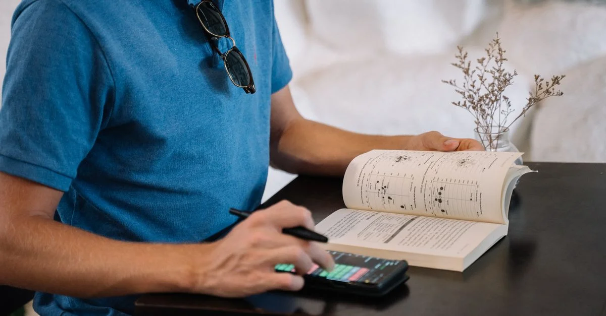 Un homme adulte utilisant un smartphone et lisant un livre à une table à l'intérieur.