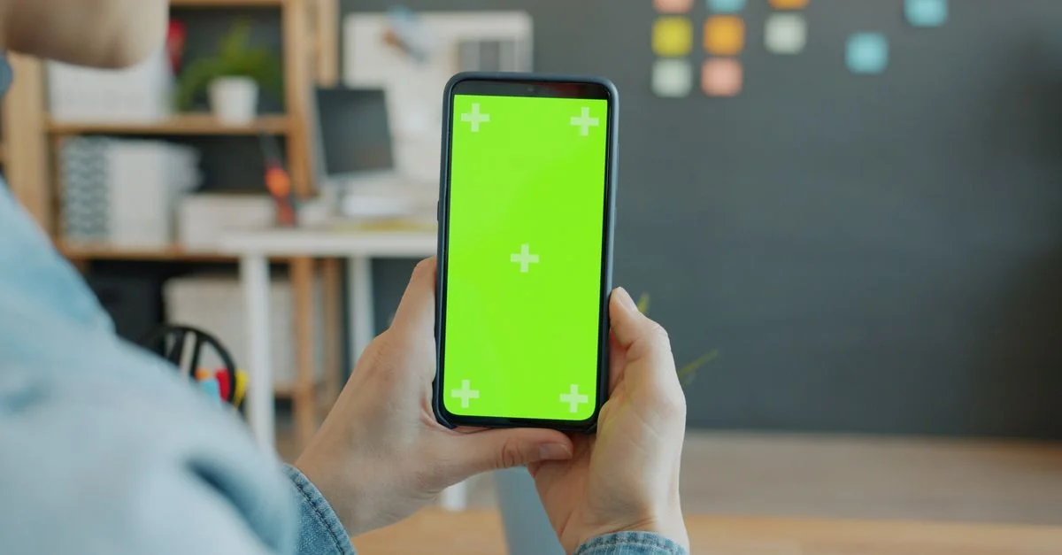 Des mains tenant un smartphone à écran vert dans un environnement de bureau moderne.