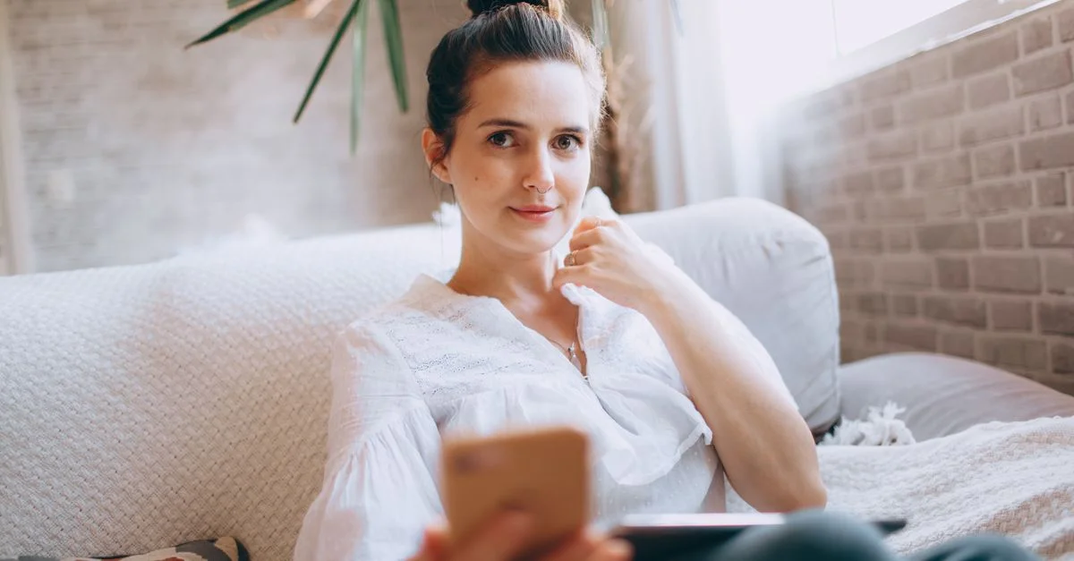 Jeune femme tenant un smartphone à l'intérieur, se relaxant sur un canapé dans un salon lumineux.